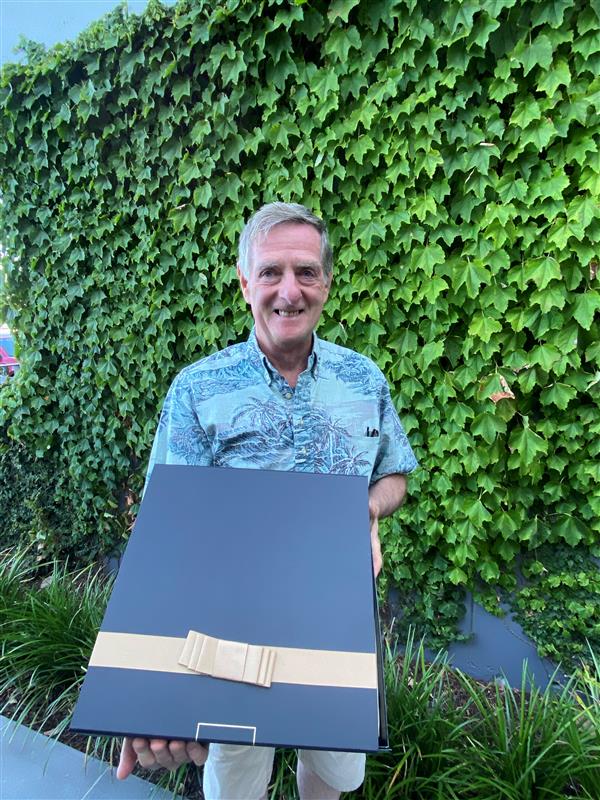 Man holding a black box iwt a gold ribbon and bow standing infront of a wall covered with ivy.